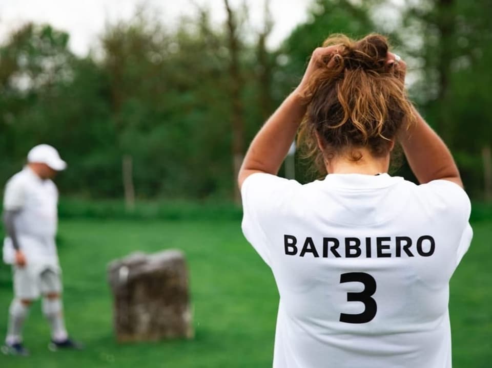 Frau mit 'Barbiero 3' auf ihrem Shirt schaut auf eine Golfspielerin im Freien.