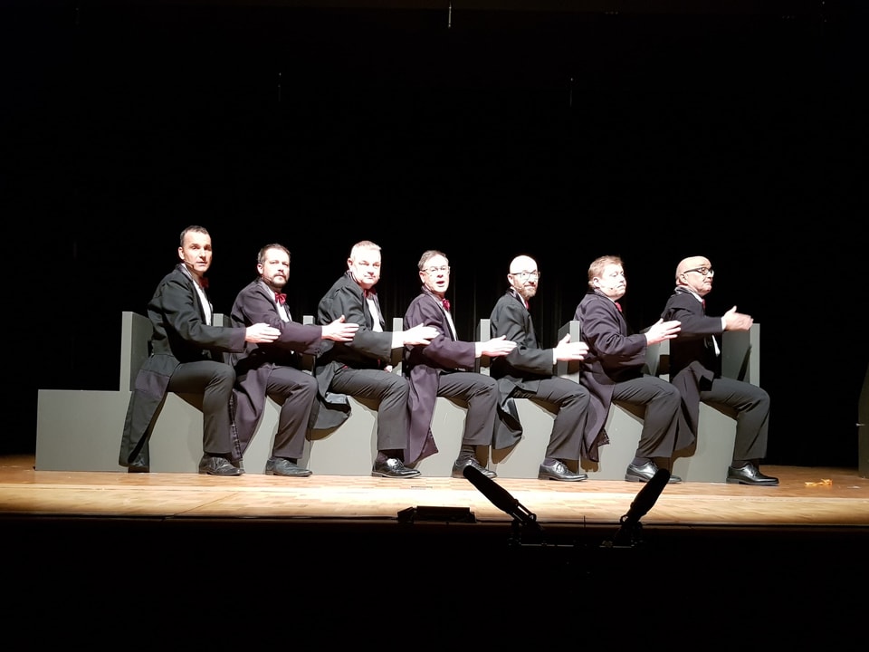 Sieben Männer im schwarzen Anzug sitzen hintereinander und singen.