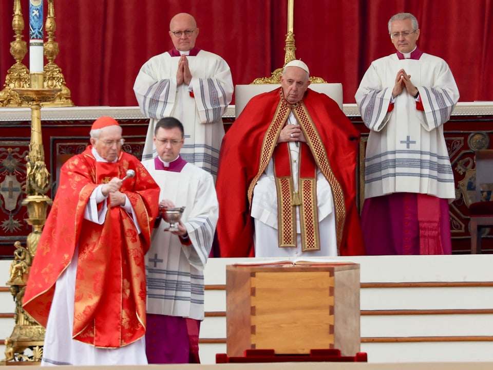 Papst Franziskus blickt sitzend von einem Podium auf den vor ihm aufgestellten Sarg herab, der von einem weiteren Geistlichen gesegnet wird.