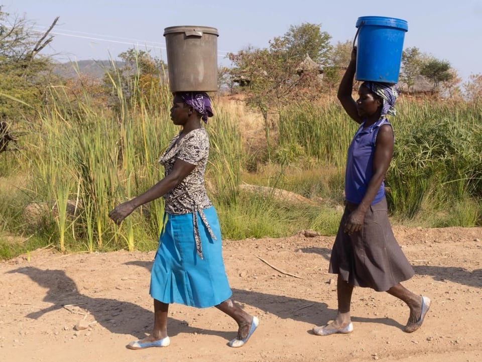 Zwei Bäuerinnen laufen über einen staubigen Weg und tragen grosse Plastikkübel mit Wasser auf dem Kopf.