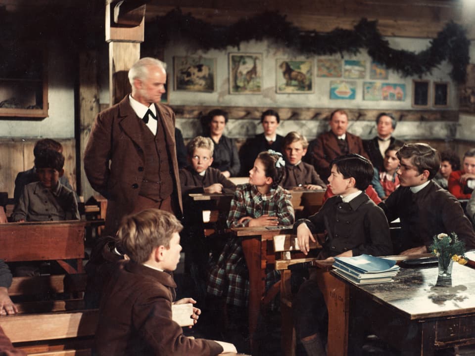 Im Klassenzimmer. Heidi und andere Schüler sitzen in den Schulbänken. Der Lehrer steht neben Heidi und schaut auf sie hinab.