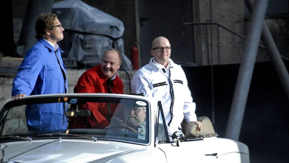 Drei Männer in farbigen Overalls in einem offenen, weissen Wagen