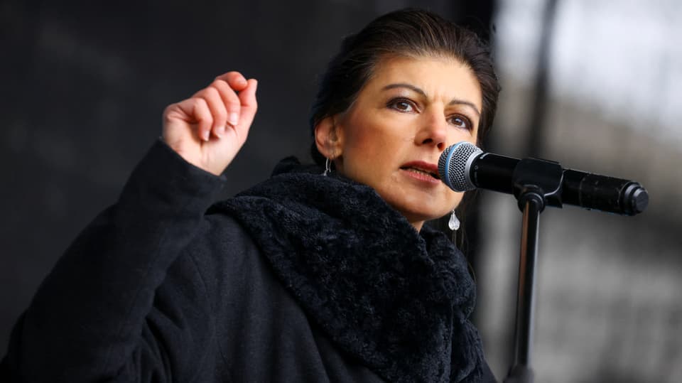 Sahra Wagenknecht spricht an einem Mikrophon und streckt die rechte Hand in die Höhe.