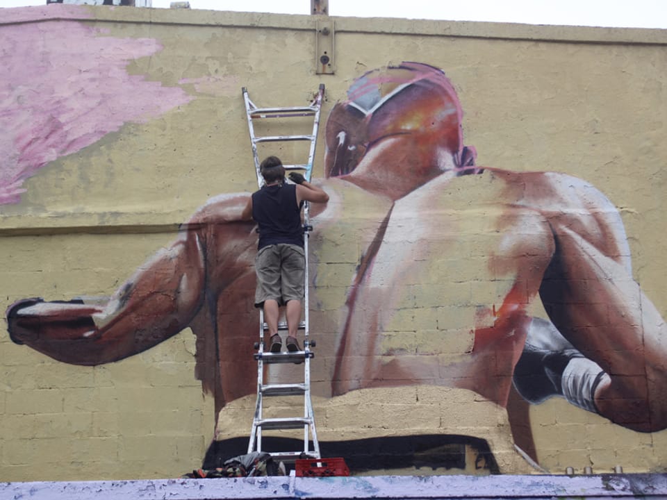 Mann auf Leiter sprayt ein Bild eines Boxers an eine Wand.