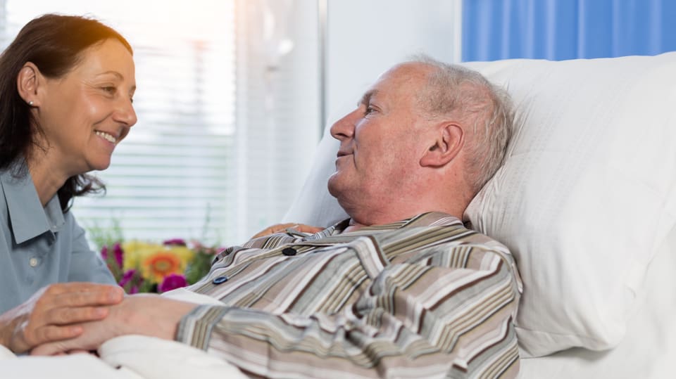 Ein alter Mann liegt im Krankenbett, eine jüngere Frau hält seine Hand.