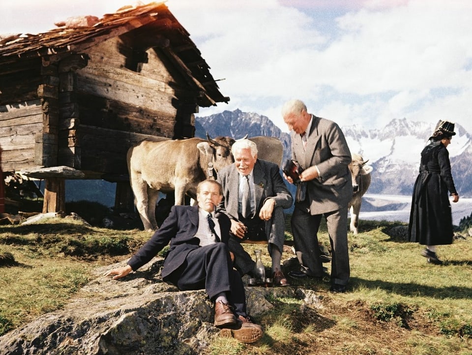 Drei Männer im Anzug auf einer Alm. Einer sitzt auf einem Stein, der zweite auf einem Melkstuhl, der dritte steht daneben. Hinter ihnen steht eine Alphütte.