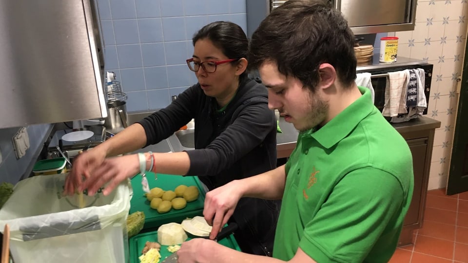 Ein Mann in einem grünen T-Shirt und eine Frau mit schwarzem T-Shirt schneiden Gemüse in einer Küche.