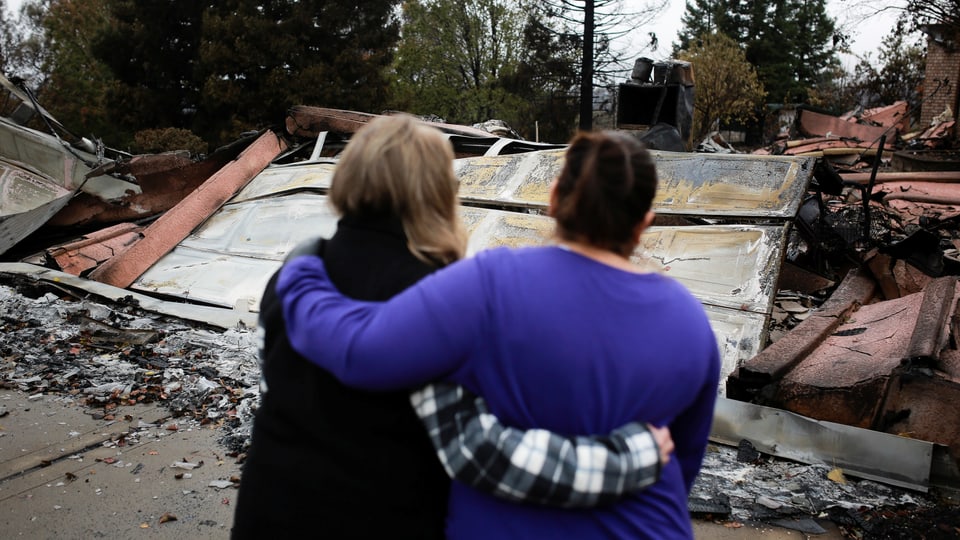Zwei Frauen stehen Arm in Arm vor ihrem zerstörten Hab und Gut.