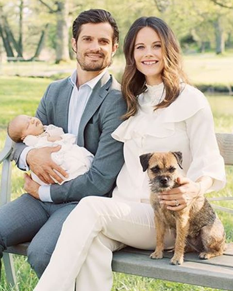 Carl-Philip ein Baby in den Armen haltend. Er sitzt zusammen mit seiner Frau und einem Hund auf einer Bank in einem Park.