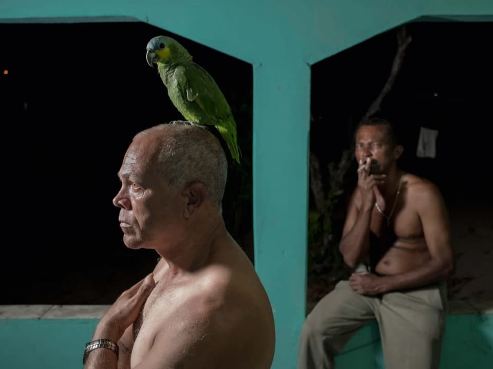 Zwei ältere Männer mit nacktem Oberkörper: einer hat einen grünen Papagei auf dem Kopf, der andere raucht.
