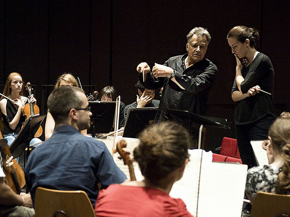 Der Dirigent umbegen von Musikerinnen und Musikern an einer Probe.