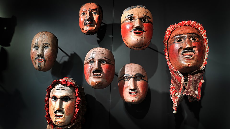 An einer schwarzen Wand hängen sieben männliche Masken.