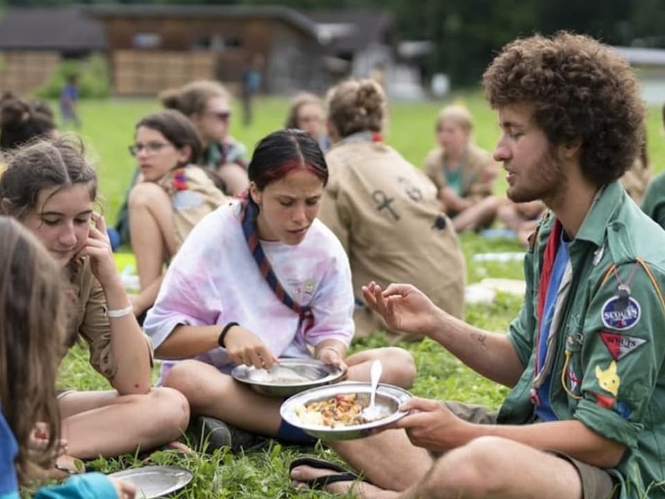 Pfadileiter sitzt mit Teilnehmerinnen auf der Wiese und isst.