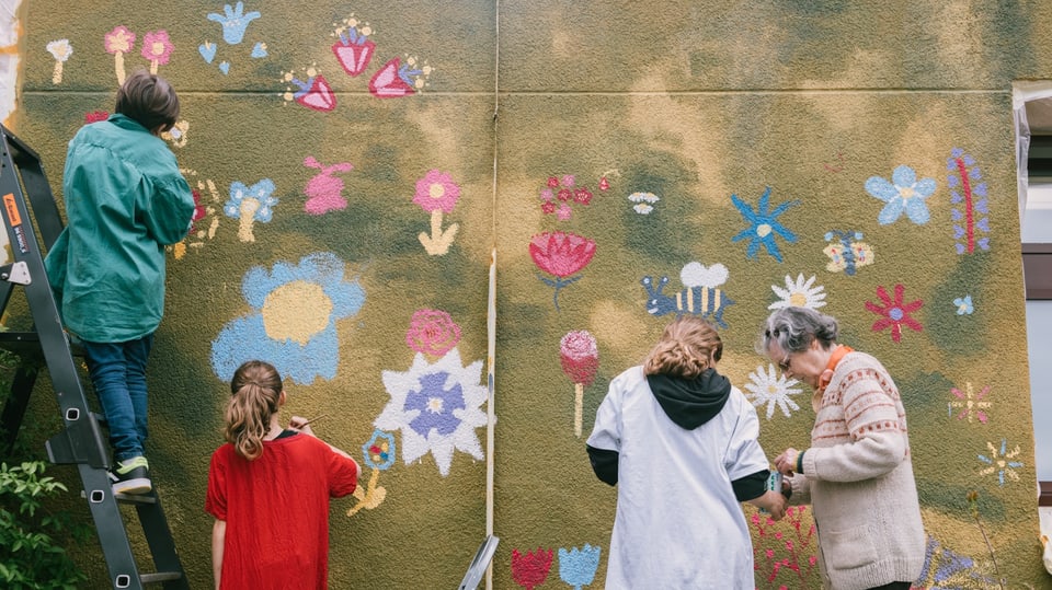 Kinder, Jugendliche und eine Senioren malen Blumen auf eine Fassade.