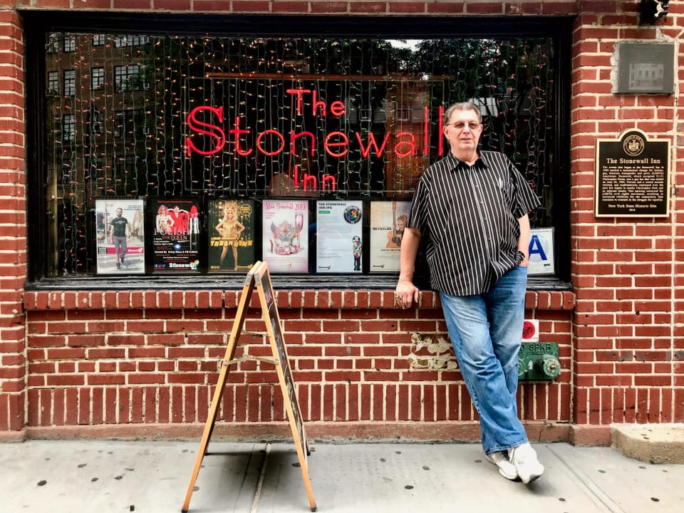 Ein Mann mit gestreiftem Hemd und Jeans steht vor einer Bar. Im Schaufenster ist eine rote Leuchtschrift "The Stonewall Inn".