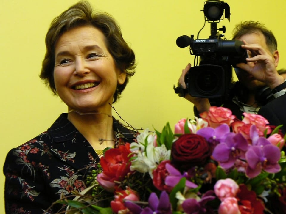 Elisabeth Kopp lächelt und trägt einen Blumenstrauss, während ein Journalist sie filmt.