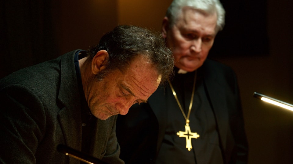 Ein Mann und ein Pfarrer mit goldenem Kreuz um den Hals. Beide in Schwarz gekleidet.
