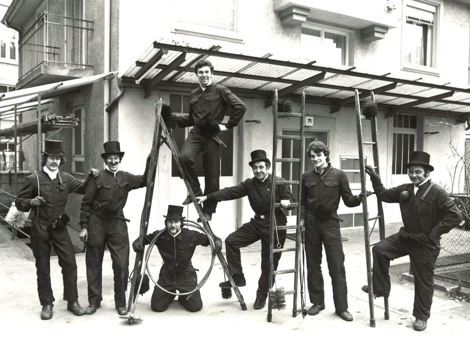 Schwarz-Weiss-Fotografie mit sieben Kaminfegern mit Leitern und Reinigungsutensilien.