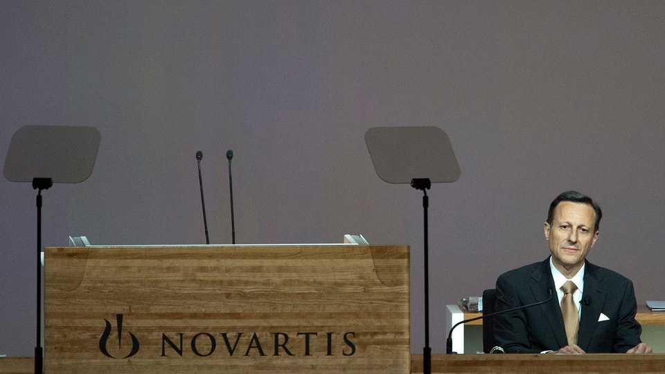 Zu sehen ist ein mit edlen Holzpanelen verkleidetes Rednerpult mit zwei Mikrophonen. Das Rednerpult ist mit dem Schriftzug Novartis angeschrieben. Das Rednerpult ist verwaist. Daneben sitzt Daniel Vasella in Anzug und Kravatte und neutralem Gesichtsausdruck.