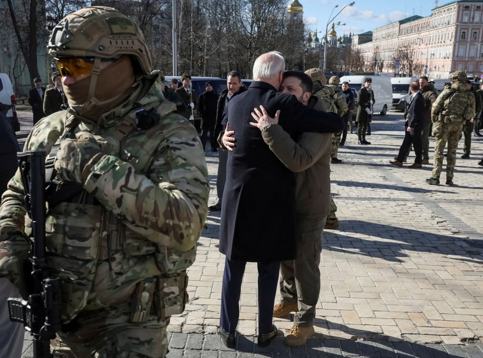 Zwei Männer umarmen sich auf einer Strasse. Im Vordergrund steht ein schwer-bewaffneter Soldat in Helm und Uniform.