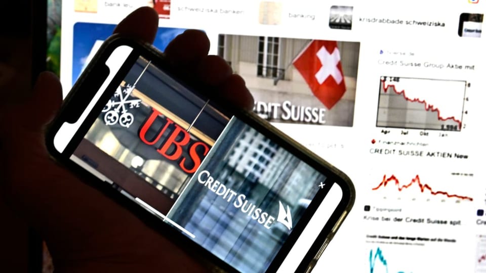 Eine rechte Hand hält ein Smartphone schräg; auf dem Bildschirm ist das Logo von UBS und Credit Suisse zu sehen.