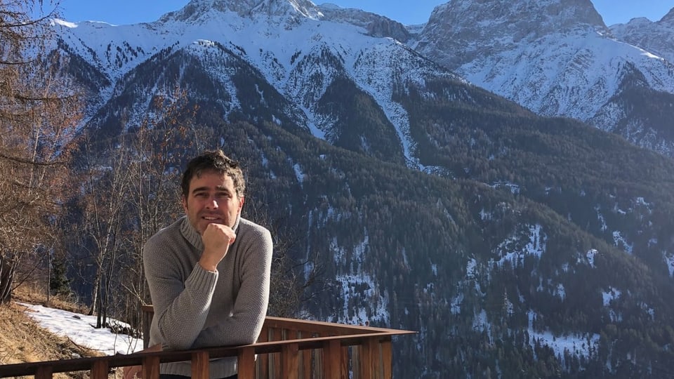 Mann mit dunklem Haar und grauem Pulli lehnt an Holzbalkon, dahinter die Berge und blauer Himmel