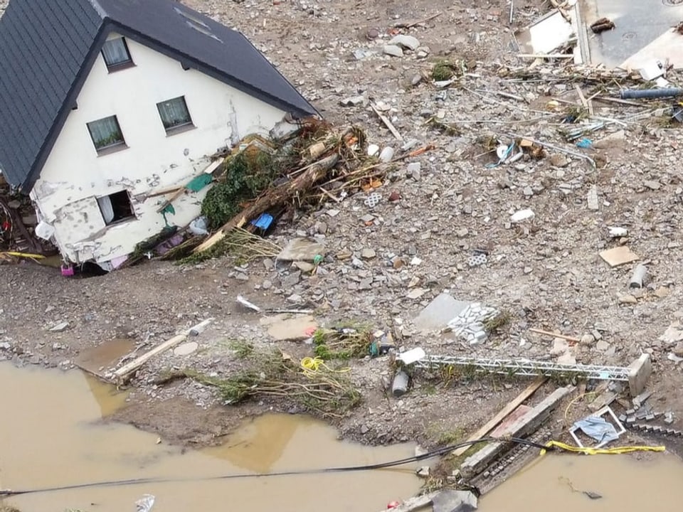 Ein völlig zerstörtes, schiefes Haus, umgeben von Schutt und Wasser.