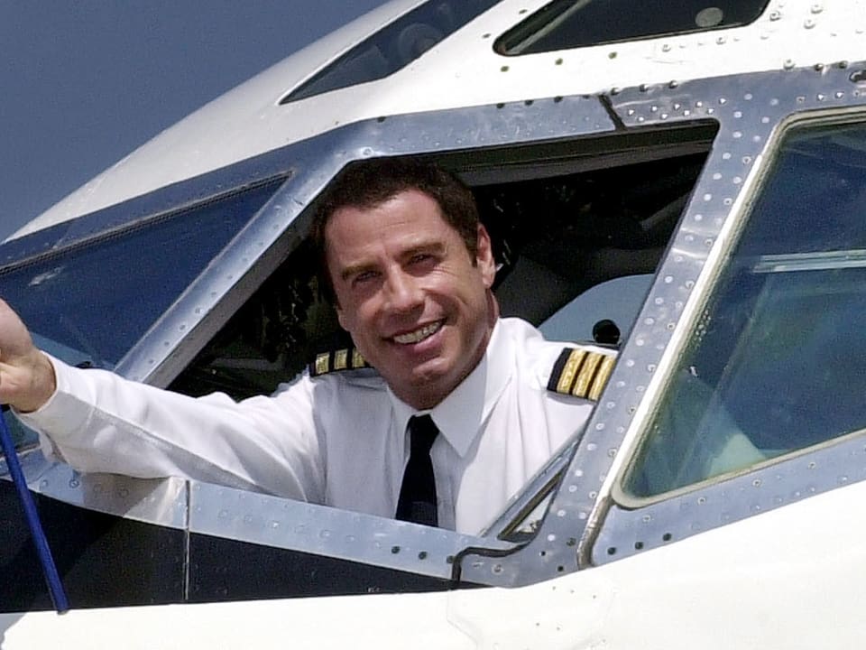 John Travolta winkt aus einem Flugzeug-Cockpit-Fenster.