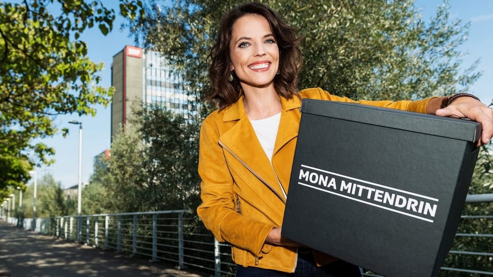 Mona Vetsch steht vor dem SRF-Fernsehstudio und hält die Blackbox mit der Aufschrift «Mona mittendrin» unter dem Arm.