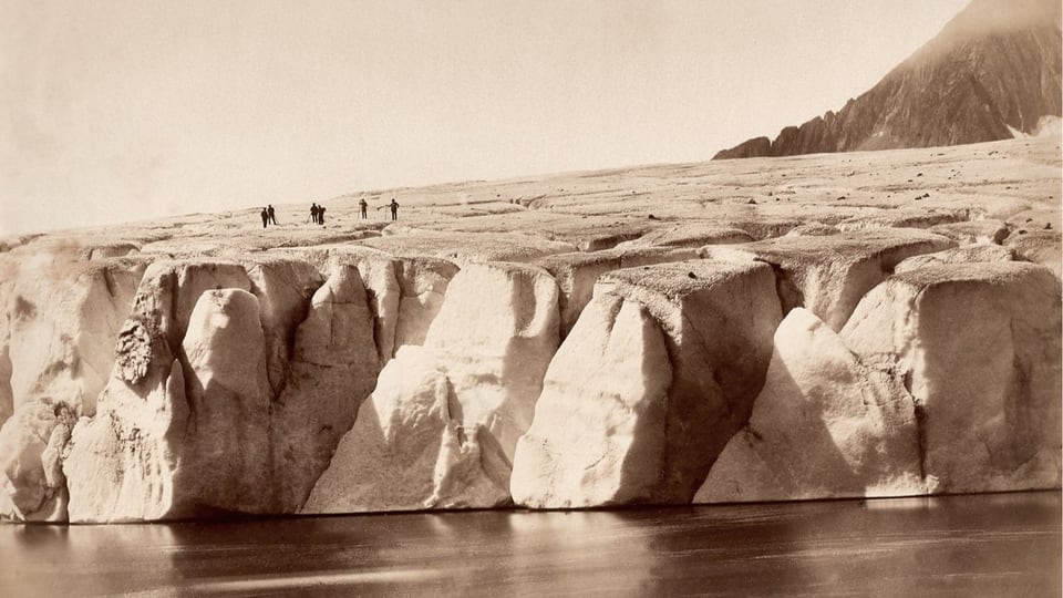 schwarzweiss Foto, vorne See, hinten riesige Gletschermasse, darauf ganz klein einige Menschen.