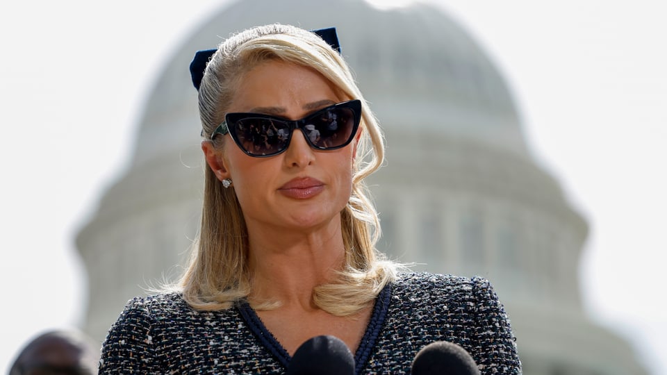 Paris Hilton steht vor einem Mikrofon und wirkt bedrückt. Sie setzt sich für mehr Kindeswohl in Erziehungsheimen ein.
