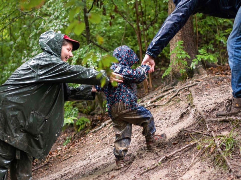 Zwei Erwachsene in Regenkleidung ziehen und schieben ein Kleinkind in Regenkleidung einen matschigen Hang hinauf.