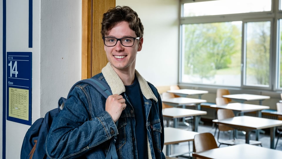 Ein Jugendlicher steht vor einem Klassenzimmer und lächelt in die Kamera.