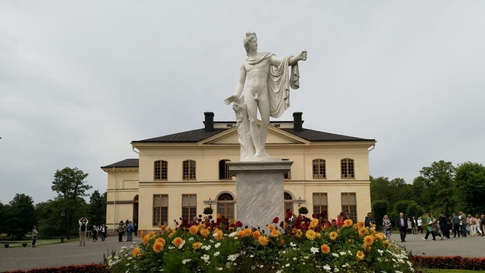 Das «Drottningholms Slottsteater» Eine weisse Statue, umgeben von Blumen, vor einem dreistöckigem grossen Gebäude.