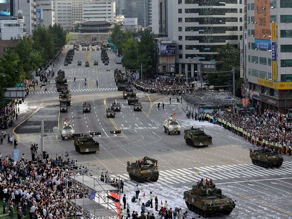 Panzer befahren eine breite Strasse in Seoul, Menschenmassen stehen an den Strassen und beobachten die Parade. Im Hintergrund dieser Aufnahme sind Hochhäuser zu sehen. 