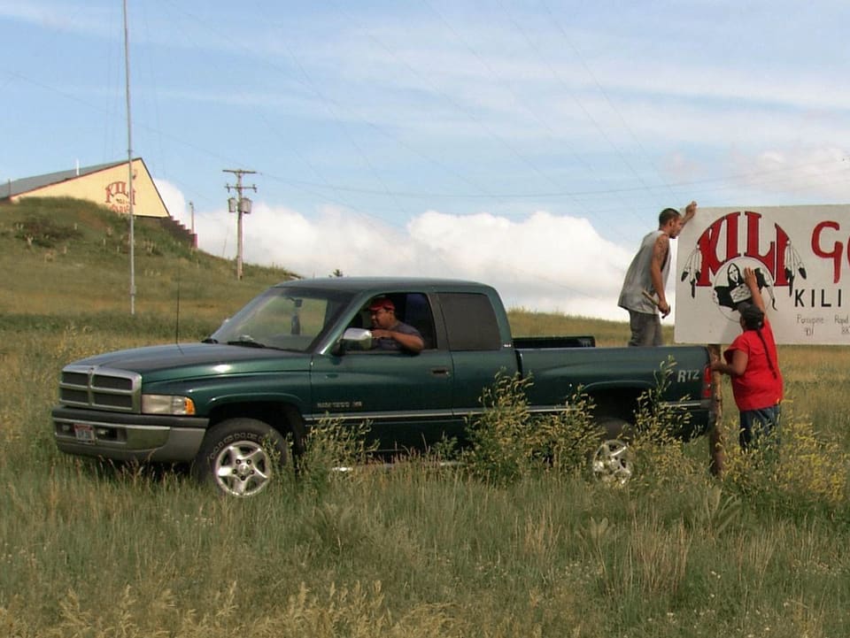 Filmstill: Ein Pickup-Truck und junge Männer, die ein Schild aufstellen.