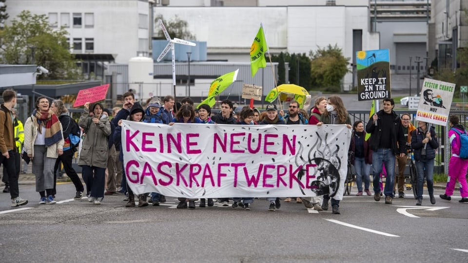 Ein Demonstrationszug läuft auf Strasse. Leute halten Fahnen und Plakate hoch, auf einem steht: Keine neuen Kraftwerke