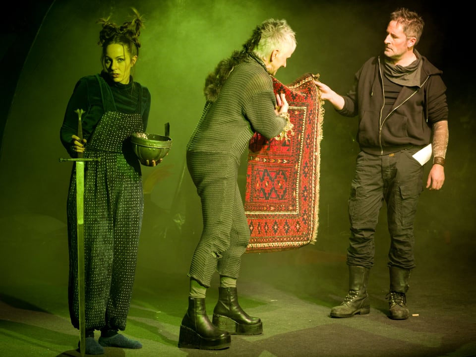 Drei Schauspiele auf einer Bühne. Sie halten ein Schwert und einen Teppich in den Händen
