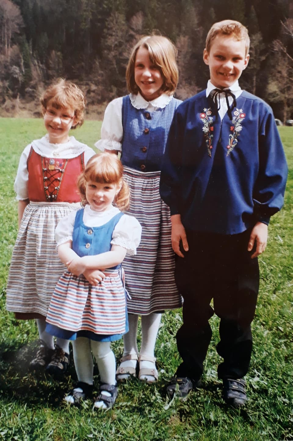 Vier Kinder. Ein Junge der eine blaue Tracht trägt. Die Mädchen tragen rote und blaue Trachten mit 