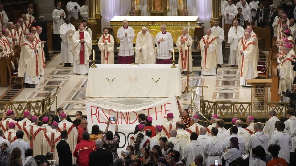 Der Papst wird während einer Messe aufgefordert, die Doktrion der Entdeckung zu widerrufen. Ein Plakat in einer Kirche