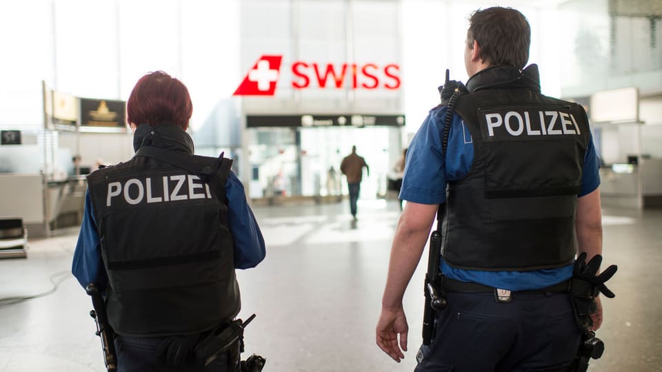 Zwei Polizisten von hinten fotografiert patrouillieren in einer Halle am Flughafen Zürich, zwischen ihnen im Hintergrund der Schriftzug "Siwss"