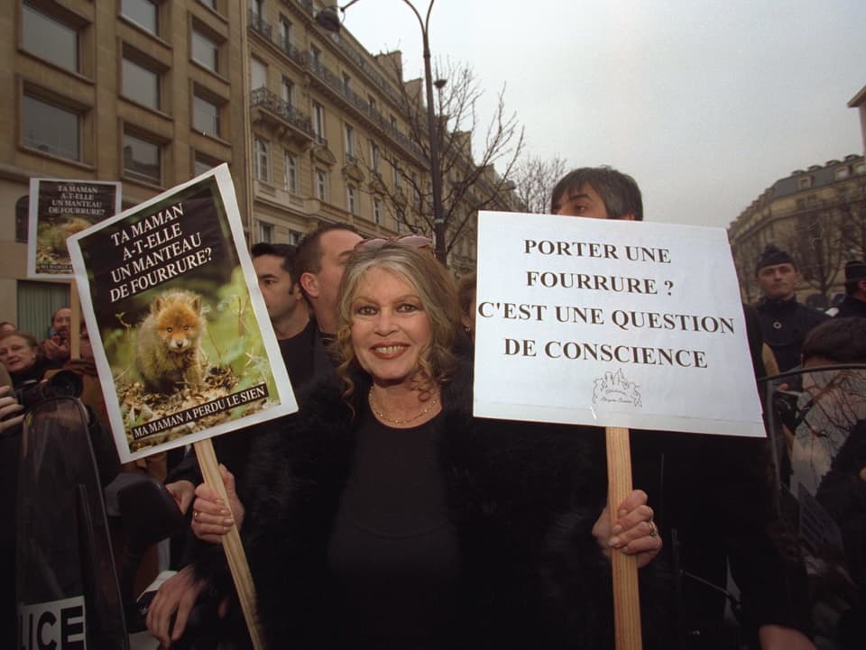 Brigitte Bardot an einer Demonstration gegen Tierpelz mit zwei Plakaten in der Hand. 