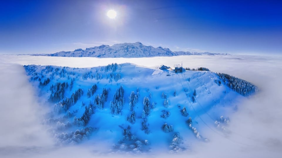 Winterfoto mit Nebelmeer. Im Vordergrund ragt ein Berg mit Bergbahn und Bäumen aus dem Nebel. 