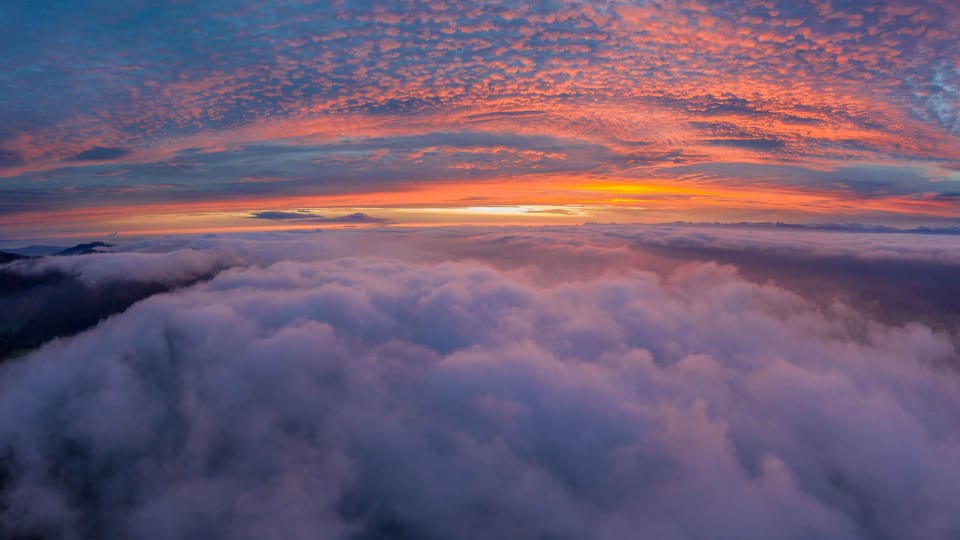 Drohnenaufnahme: Oben Wolkenfelder und Morgenrot, unten aufgelockerter Nebel.