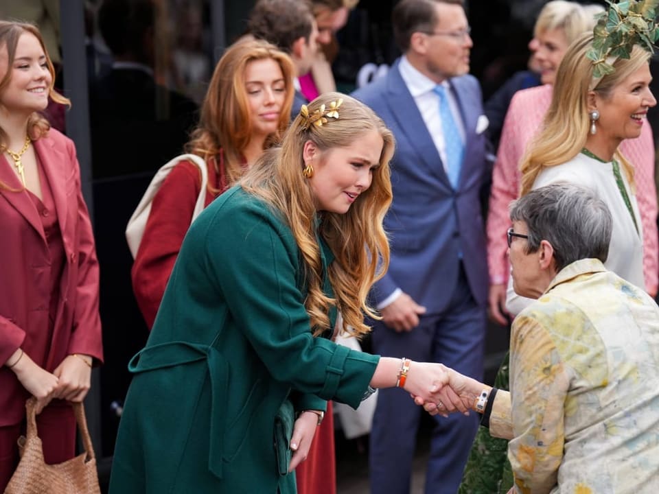 Frau in grünem Mantel begrüsst ältere Dame mit Handshake bei gesellschaftlichem Ereignis