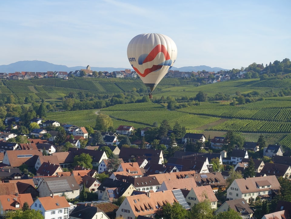Anlässlich des 90 jährigen Jubiläums hat die Ballongruppe Basel eine Fahrt mit 7 Ballonen über die Stadt gemacht. Von Schönenbuch über die Stadt bis nach Weil am Rhein in Deutschland. Man sieht einen Heissluftballon über den Dächern von Schönenbuch. Dahinter Weinreben.