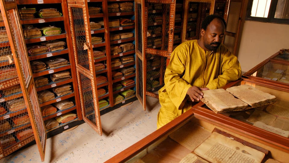 Ein schwarzer Mann in gelbem Kleid sitzt in einer Bibliothek, vor ihm ein altes Manuskript.