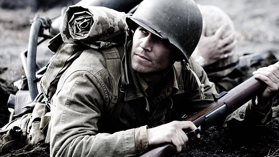Ein junger Soldat mit Helm und Gewehr liegt am Boden und schaut kritisch.