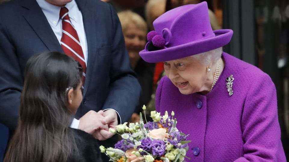 Eine lachende Queen im violettem Mantel. Eine Mädchen überreicht ihr einen Blumenstrausse mit verschiedenen Blumen.
