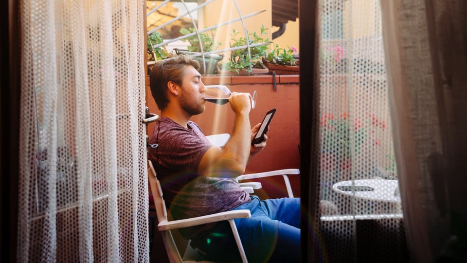 Ein Mann sitzt am Balkon mit E-Reader und trinkt ein Glas Wein.